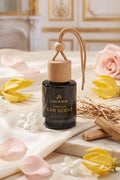 A bottle of Akasia Premium Car Scent with a natural wood cap and hanging loop, styled on a marble surface with pink rose petals, yellow tulips, and dried botanicals in a warm, elegant interior setting.