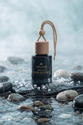 A car fragrance bottle labeled AKASIA PREMIUM CAR SCENT, with a dark glass body, light wood cap, and hanging cord, placed among stones in shallow water with a misty blue background.